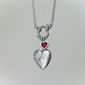 May include: A silver chain necklace with a heart-shaped locket and a smaller red heart charm. The locket has a detailed design and the charm has a red gemstone.