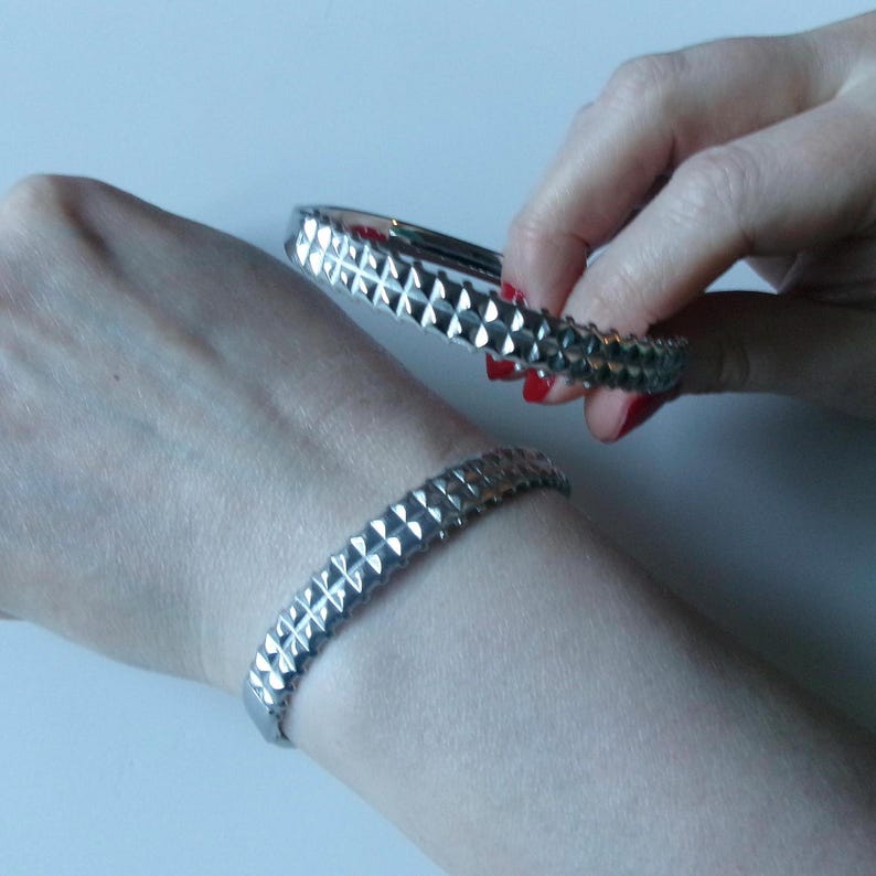 May include: Two silver-colored, studded bracelets. One bracelet is on a wrist, and the other is held by a hand. The bracelets have a geometric design with raised, pyramid-shaped studs. The background is a plain, light surface.