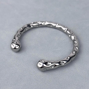 May include: A silver-toned metal cuff bracelet featuring an open design and a textured, uneven surface with multiple small, rounded indentations along the entire length of the band. The ends of the bracelet are rounded and smooth in appearance.