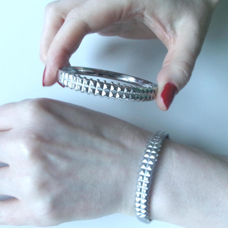 May include: Silver-colored bangle bracelet with a repeating geometric pattern. The bracelet is shown held in a hand with red nail polish, and also worn on a wrist. The design features a series of raised, pyramid-shaped elements.