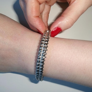May include: Silver-toned bracelet with a repeating pattern of pyramid-shaped studs. The bracelet is shown on a wrist, with a hand holding it. The bracelet appears to be made of metal.