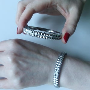 May include: A silver bangle bracelet featuring a repeating pattern of raised, pyramid-shaped studs. The bracelet is held above another similar bracelet worn on a wrist. The bracelets are made of a shiny metal.