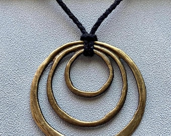 May include: A bronze-toned pendant necklace featuring three nested, irregularly shaped rings. The rings are suspended from a black cord, creating a layered, circular design. The necklace has a rustic, handcrafted appearance.