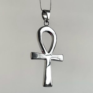 May include: A silver-colored Ankh pendant necklace. The pendant is a stylized cross with a loop at the top, suspended from a thin chain. The Ankh is a symbol of life and immortality.