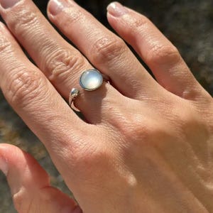 May include: A silver ring with a round, iridescent, light-colored stone. The ring has an open design, with the stone set on top of the finger. The band is smooth and shiny, and the stone appears to be a cabochon.