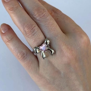 May include: Silver-toned ring featuring a bow design with a pink heart-shaped center stone. The ring is worn on a finger, showcasing the intricate details of the bow and the sparkle of the gemstone.