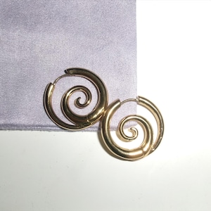 May include: A pair of gold-colored spiral hoop earrings. The earrings have a circular design with a spiral detail in the center. The earrings are resting on a light purple fabric.
