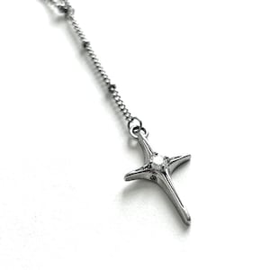 May include: A silver-colored pendant necklace featuring a stylized cross design. The cross has four elongated points and a small, round accent at the center. The necklace chain is a delicate, beaded style.