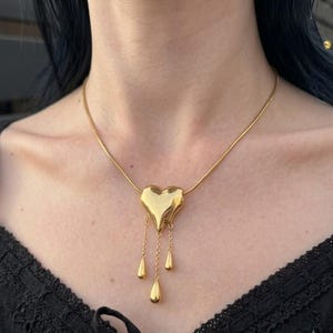 May include: A gold-toned necklace featuring a smooth, puffed heart pendant. Three delicate gold chains hang from the bottom of the heart, each ending in a small, polished gold teardrop-shaped charm. The pendant is suspended on a thin gold snake chain.