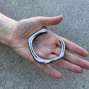 Pode incluir: Uma pulseira de punho prateada polida com um design quadrado e aberto. A pulseira é segurada em uma mão, mostrando sua superfície lisa e reflexiva. Uma peça de joalheria moderna.