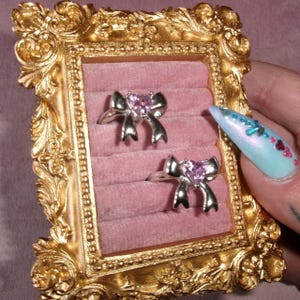 May include: Two silver rings with bow-shaped designs, each featuring a pink heart-shaped stone. The rings are displayed in a pink velvet-lined box within a gold ornate frame. The frame is held by a hand with a long, light blue nail.