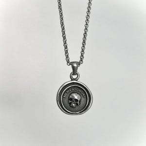 Op de afbeelding: Een zilverkleurige metalen ketting met een ronde hanger met een verhoogd schedelontwerp in het midden. De tekst MEMENTO MORI is in een cirkelvormig patroon rond de schedel gegraveerd. De hanger hangt aan een bijpassende zilverkleurige schakelketting.