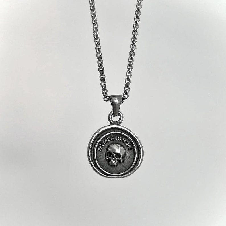 Op de afbeelding: Een zilverkleurige metalen ketting met een ronde hanger met een verhoogd schedelontwerp in het midden. De tekst MEMENTO MORI is in een cirkelvormig patroon rond de schedel gegraveerd. De hanger hangt aan een bijpassende zilverkleurige schakelketting.