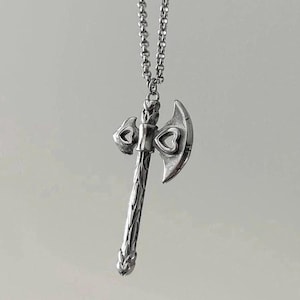 May include: A silver necklace with a detailed axe pendant. The axe has a heart-shaped design on the blade and a textured handle.