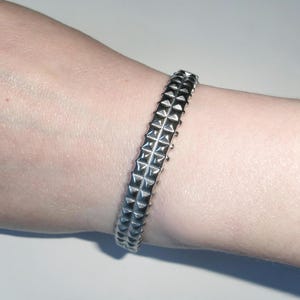 May include: A silver-colored bracelet with a studded design. The bracelet is worn on a wrist and features rows of pyramid-shaped studs. The metal has a shiny, reflective surface, and the bracelet appears to be made of a solid material.