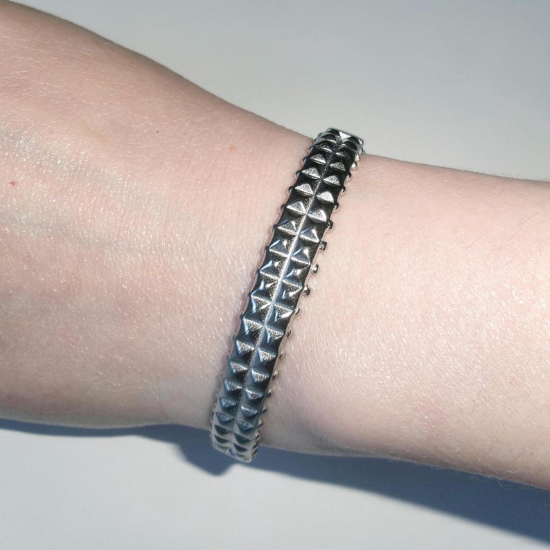 May include: A silver-colored bracelet with a studded design. The bracelet is worn on a wrist and features rows of pyramid-shaped studs. The metal has a shiny, reflective surface, and the bracelet appears to be made of a solid material.