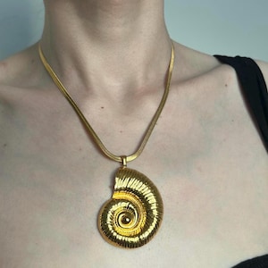 May include: A gold-toned necklace featuring a large, spiral pendant resembling a seashell. The pendant is intricately detailed with textured lines and a polished finish. The chain is a delicate, flat design, creating a striking contrast with the bold pendant.