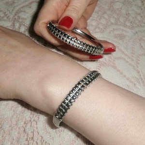 May include: Silver-toned metal bracelets with a studded design. One bracelet is worn on a wrist, while another is held in a hand with red nail polish. The bracelets have a geometric pattern.