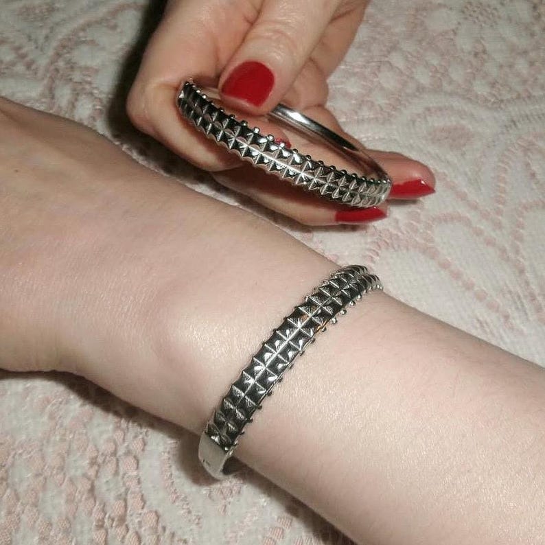 May include: Silver-toned metal bracelets with a studded design. One bracelet is worn on a wrist, while another is held in a hand with red nail polish. The bracelets have a geometric pattern.