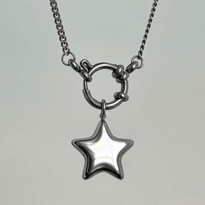 Op de afbeelding: Een zilverkleurige ketting met een sterhanger. De ketting heeft een dikke ketting en een grote ronde sluiting. De sterhanger heeft een vijfhoekige vorm met een gepolijst, reflecterend oppervlak.