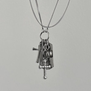 May include: A silver-toned necklace with multiple charms. The necklace features a dog tag, two crosses, and other small charms. The chain is a delicate, layered design. The pendant hangs from a circular ring.