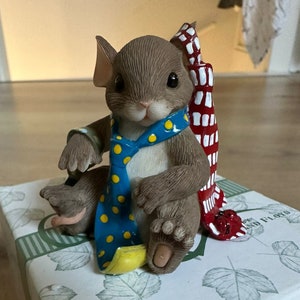 Op de afbeelding: Een klein, bruin muisbeeldje dat een blauwe en witte stippen stropdas en een rood-witte gestreepte sjaal draagt. Het beeldje zit op een witte doos met een groen bladmotief.