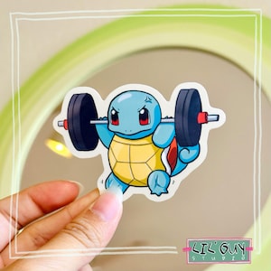 May include: A sticker featuring a cartoon Squirtle character lifting a barbell. The Squirtle is blue with a yellow shell and red accents. The barbell has black weights and red ends. The sticker has the text "LIL' GUY STUDIO".