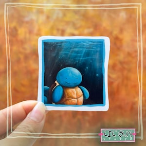 May include: A sticker depicting a blue cartoon turtle with a smiling expression, set against a blue underwater scene. The turtle has a tan shell and blue limbs. The sticker is held by a hand, with the text "LIL' GUY STUDIO" at the bottom.