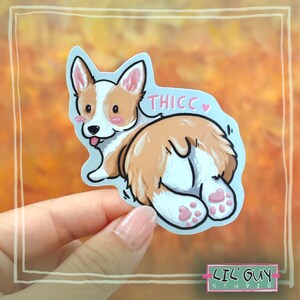 May include: A sticker of a cartoon corgi with a pink heart and the text "THICC" on a blue background. The corgi is brown and white with a pink tongue sticking out. The sticker is held in a hand.