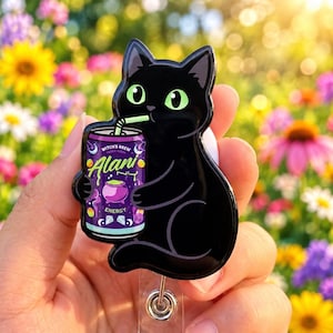 May include: A black cat badge reel featuring a glossy finish and bright green eyes. The cat is holding a purple Alani Nu energy drink can labeled Witch's Brew with a graphic of a cauldron, stars, and crystals, complete with a light green straw.