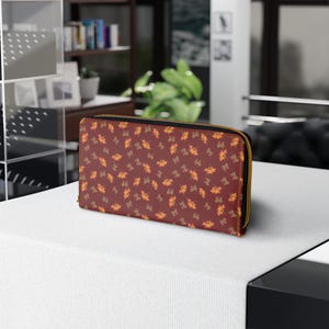 May include: A burgundy wallet with a gold zipper, featuring a repeating pattern of autumn leaves in shades of orange and brown. The wallet is rectangular and appears to be made of a textured material.
