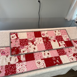 May include: A set of Valentine's Day themed quilted table runners and placemats. The patchwork design features red, pink, and white squares with heart and love-themed patterns. The edges are finished with a gray border.