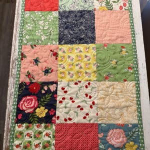 May include: A quilted table runner with a patchwork design. The runner features squares of various floral patterns in red, green, pink, yellow, and navy blue. The edges are trimmed with a green polka dot fabric.