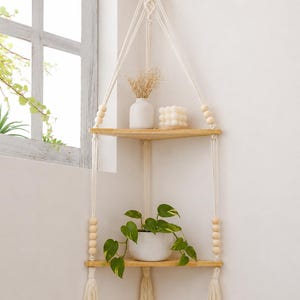 May include: A two-tier corner hanging shelf featuring light wood triangular boards suspended by cream-colored macrame cord. The structure includes natural wooden beads, decorative tassels at the base, and is mounted from a single wooden rod.
