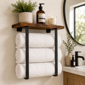 May include: A wall-mounted bathroom shelf featuring a dark brown wooden plank supported by two black metal brackets. The unit includes a lower rack holding three neatly rolled white towels, with a potted plant, soap dispenser, and candle on top.