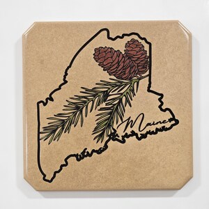 May include: A brown paper coaster with a black outline of the state of Maine. The outline includes a pine branch with pine cones in shades of brown and green. The word "Maine" is written in cursive in the lower right corner.
