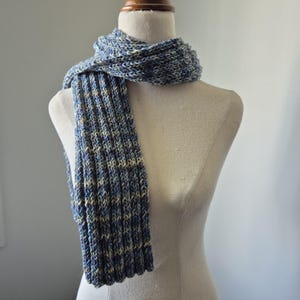 May include: A knitted scarf in shades of blue and cream, displayed on a mannequin. The scarf features a ribbed design and is made from soft yarn. The colour scheme includes blues, cream, and a hint of light brown.
