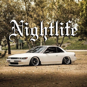 May include: A white sports car with black rims is parked on grass, with the word "Nightlife" in large, ornate white letters above it. The car is low to the ground, and the background features trees and a park setting.
