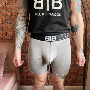 以下が含まれることがあります： 「BB BRANDON」の文字が入った黒いウエストバンドが付いたグレーのボクサーパンツ。ボクサーパンツは前面と側面がグレーで、背面には黒いパネルが付いています。モデルは「BILL & BRANDON」の文字が入った黒いタンクトップを着ています。