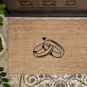 May include: A brown coir doormat with a black design of two intertwined rings and a heart above them.