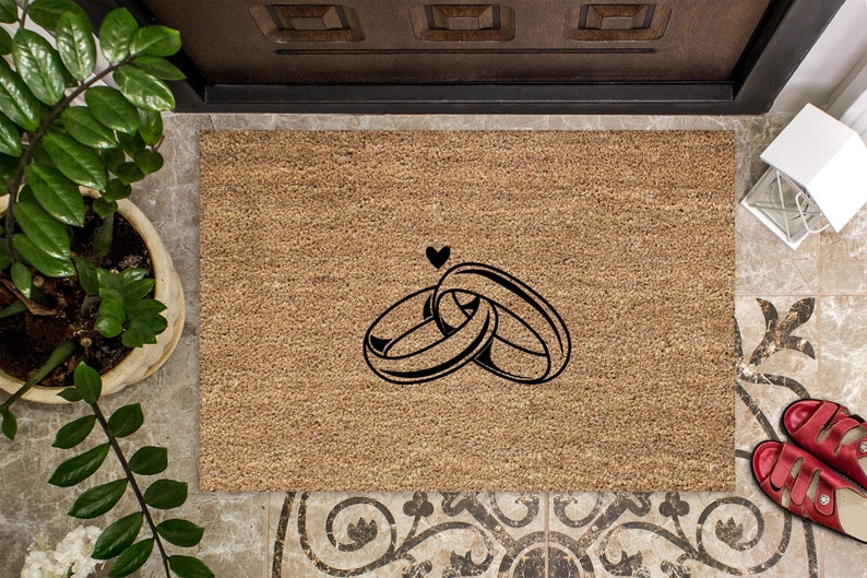 May include: A brown coir doormat with a black design of two intertwined rings and a heart above them.