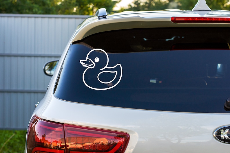 May include: A white rubber ducky decal on the back window of a silver car.