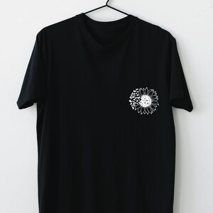 Puede incluir: Una camiseta negra con un gr&aacute;fico de girasol blanco en el pecho izquierdo. El girasol tiene un dise&ntilde;o detallado con p&eacute;talos, hojas y un centro.