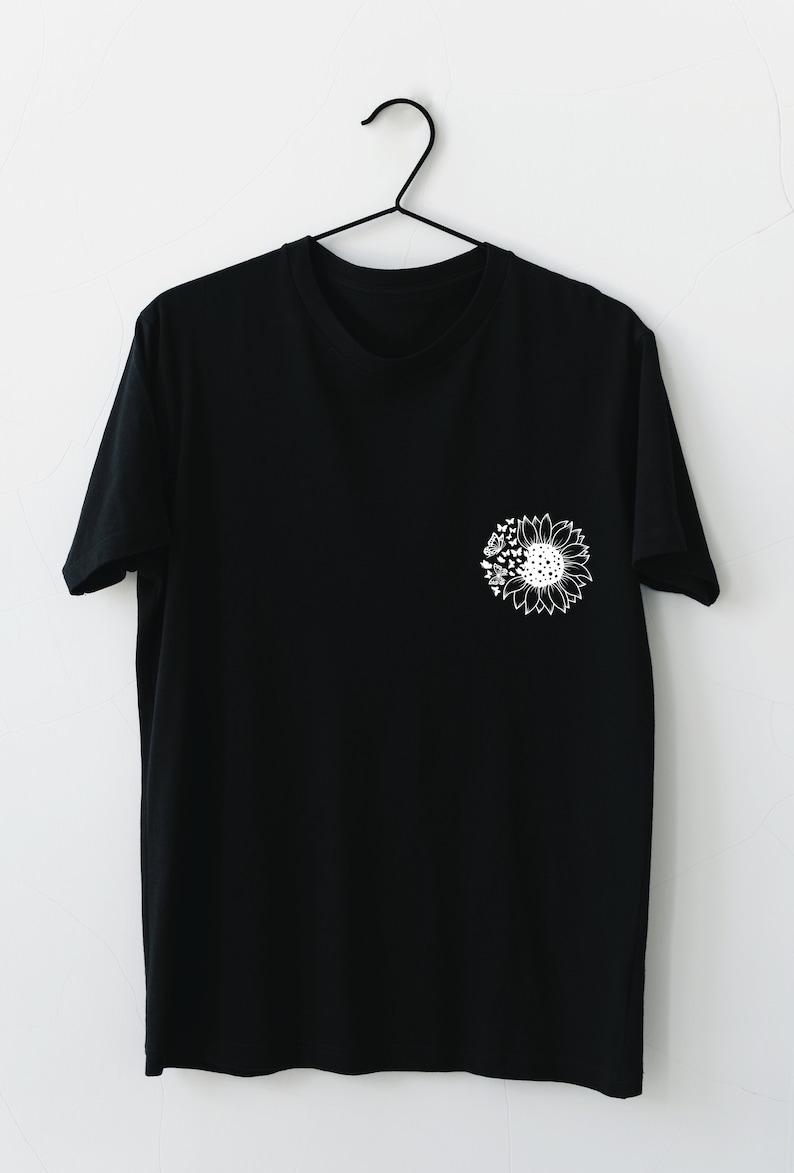 Puede incluir: Una camiseta negra con un gr&aacute;fico de girasol blanco en el pecho izquierdo. El girasol tiene un dise&ntilde;o detallado con p&eacute;talos, hojas y un centro.