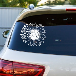 Puede incluir: Un coche blanco con una calcoman&iacute;a de girasol y mariposas en la ventana trasera. La calcoman&iacute;a es una silueta blanca de un girasol con mariposas volando a su alrededor.