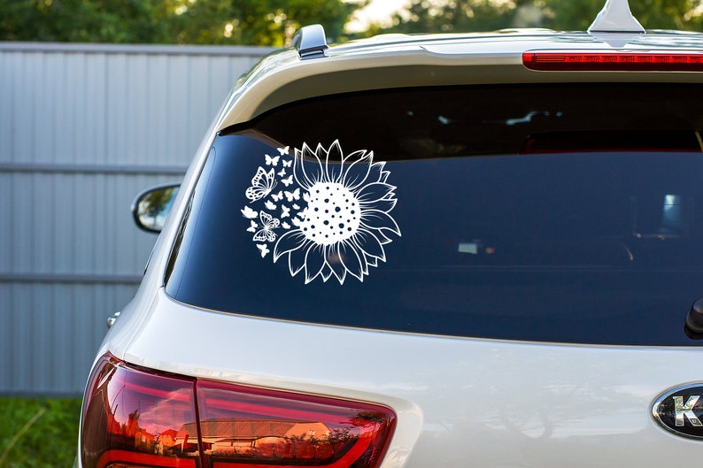 Puede incluir: Un coche blanco con una calcoman&iacute;a de girasol y mariposas en la ventana trasera. La calcoman&iacute;a es una silueta blanca de un girasol con mariposas volando a su alrededor.