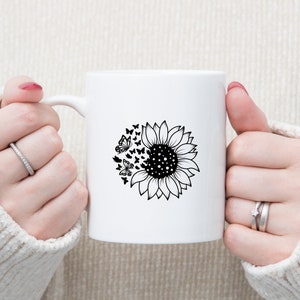 Puede incluir: Taza de cer&aacute;mica blanca con un dise&ntilde;o en blanco y negro de un girasol con mariposas que salen volando.