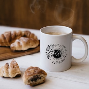 Puede incluir: Taza de cer&aacute;mica blanca con un dise&ntilde;o de girasol en blanco y negro con mariposas. La taza est&aacute; llena de caf&eacute; y sale vapor de la parte superior. Hay croissants en una tabla de cortar de madera en el fondo.