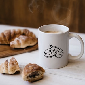 May include: White ceramic coffee mug with a black design of two interlocking rings and a small heart. The mug is filled with coffee and steam is rising from the top. There are croissants on a wooden cutting board in the background.