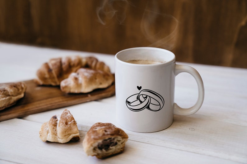 May include: White ceramic coffee mug with a black design of two interlocking rings and a small heart. The mug is filled with coffee and steam is rising from the top. There are croissants on a wooden cutting board in the background.